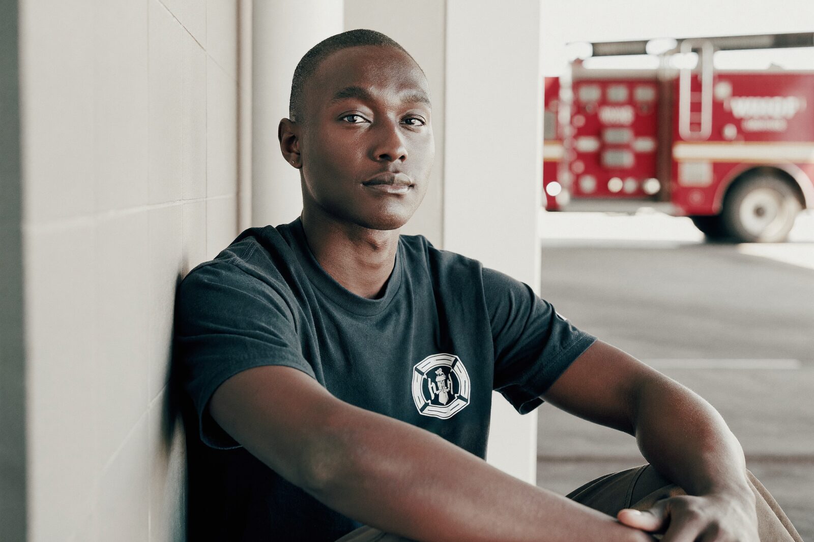 A firefighter sits in a sunlit fire-station apparatus bay, a fire engine behind him
